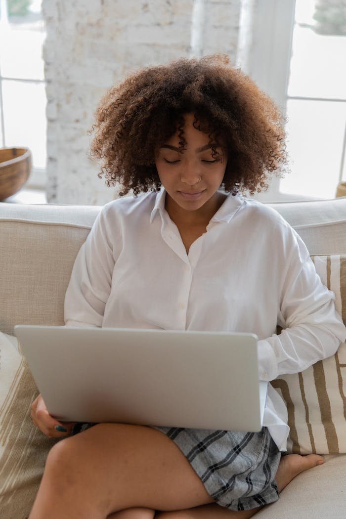 Stylish young self employed African American woman with curly hair in casual outfit sitting on comfortable sofa and working remotely on laptop in modern apartment